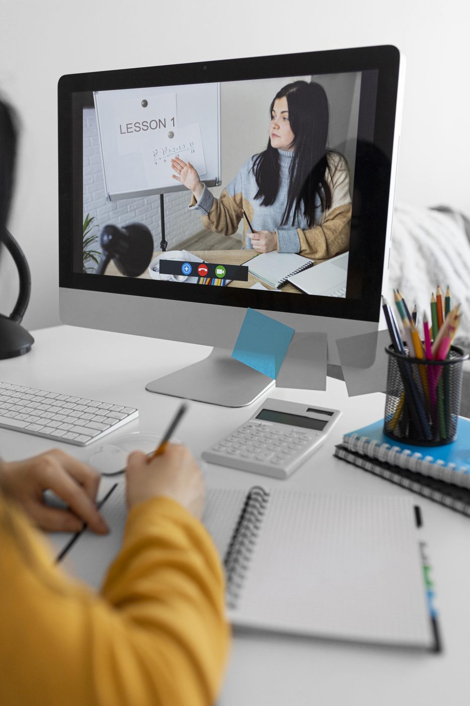 A student at a desk writing in a notebook while a tutor teaches from a whiteboard on the video call filling the screen